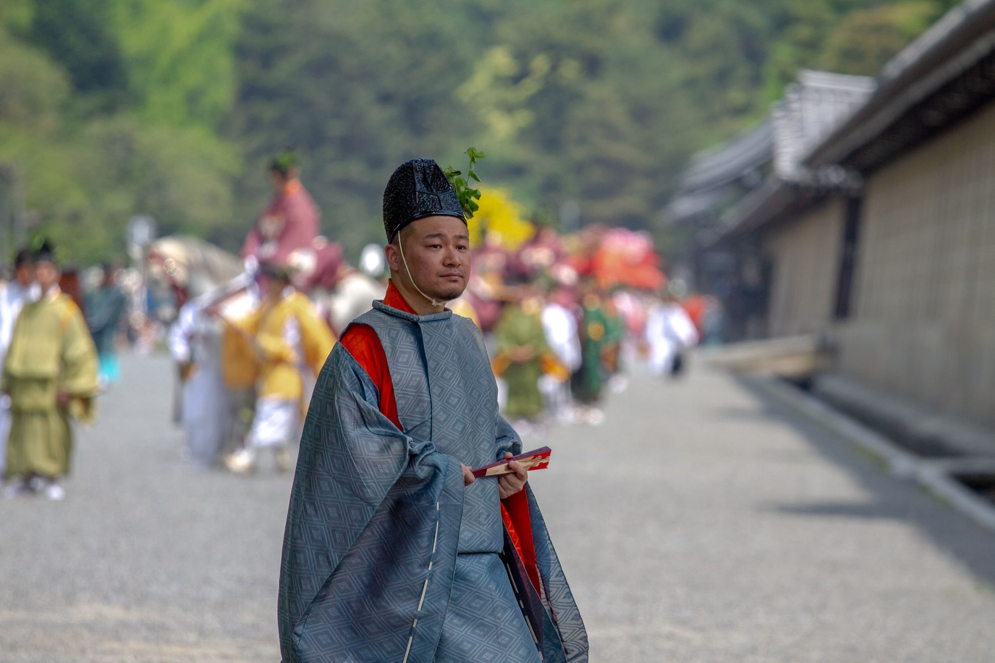 A man wearing traditional Japanese clothes. In the background out of focus is a parade procession of people in traditional styles, some mounted on horseback.