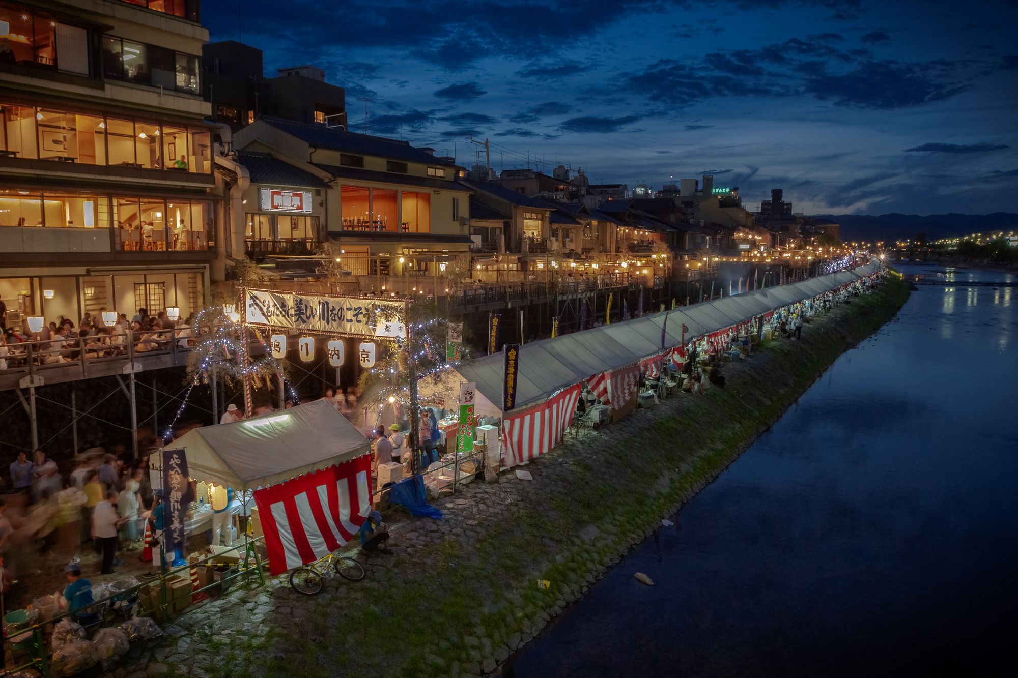 Festival tents with an inner glow along the river, with the deep blue sky of the magic hour sunset in the background, and reflected on the river water.